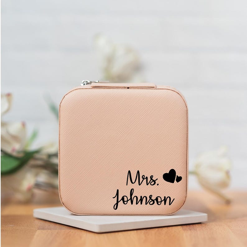 May include: A light pink jewelry box with a zipper closure. The box has the text "Mrs. Johnson" and two small hearts printed on the front.