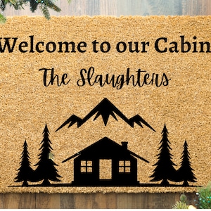 May include: Welcome to our Cabin, The Slaughters doormat with a brown background and black text. The doormat features a cabin, mountains, and pine trees.