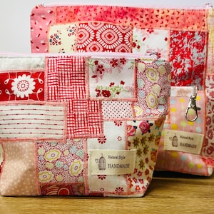 May include: Two handmade patchwork fabric pouches in red, pink, and white. The pouches feature floral and geometric patterns. One pouch has a small pocket with a metal clasp. Each pouch has a zip closure and a "Natural Style Handmade" label.