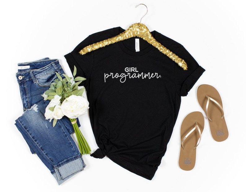 Pu&ograve; includere: T-shirt nero con la scritta "GIRL programmer" in corsivo bianco. La maglietta &egrave; esposta con jeans blu, un bouquet di fiori bianchi e un paio di infradito marroni con cinturini dorati, il tutto su sfondo bianco.