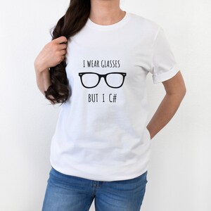 Puede incluir: Camiseta blanca con el texto negro "I wear glasses but I C#" y un gr&aacute;fico de gafas negras. La camiseta es de manga corta y cuello redondo.