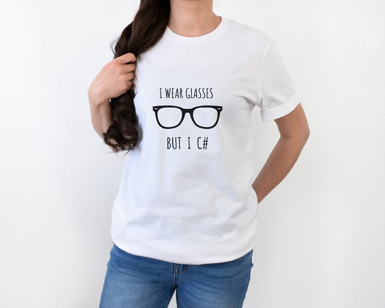 Puede incluir: Camiseta blanca con el texto negro "I wear glasses but I C#" y un gr&aacute;fico de gafas negras. La camiseta es de manga corta y cuello redondo.