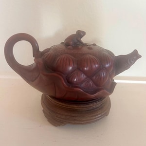 Op de afbeelding: Een donkerbruine kleitheepot met een gevormd lotusbladlichaam, een gebogen handvat en een tuit in de vorm van een vis. Het deksel is versierd met een klein kikkerfiguurtje en rust op een ronde houten standaard met een gladde afwerking.