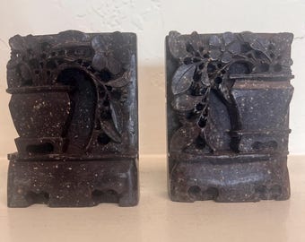 May include: A pair of dark brown, speckled stone bookends featuring intricate, carved relief designs of potted plants and floral motifs. Each rectangular piece has a solid base and a detailed, openwork top section depicting foliage.