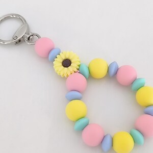 Peut inclure: Un porte-clés coloré avec un fermoir argenté. Le porte-clés présente un design de perles avec des perles roses, jaunes et bleues, et un charm en forme de tournesol jaune. Les perles mesurent environ 2 cm de diamètre.