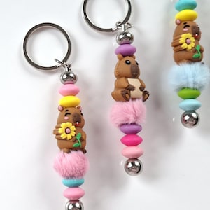 May include: Three keychains with a brown capybara charm, colorful beads, and a fluffy pom-pom. The keychains have a silver metal ring and a decorative design with pink, yellow, blue, and purple accents. The capybaras are holding a flower.