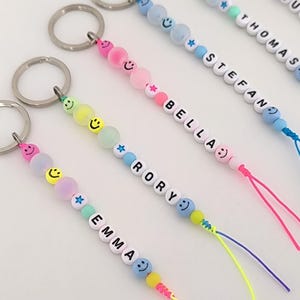 Könnte beinhalten: Eine Sammlung personalisierter Schlüsselanhänger mit silbernen Metallringen an bunten Schnüren. Jeder Anhänger ist mit runden Pastellperlen, Smiley-Perlen, sternförmigen Perlen und weißen Buchstabenperlen verziert, die Namen wie EMMA, RORY, BELLA, STEFAN, THOMAS und SUZANNE bilden.
