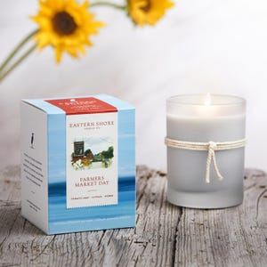 May include: A white candle with a rope tied around it, sitting on a wooden surface. The candle is lit and the flame is visible. The candle is in front of a box with a blue and white design. The box has the text "Eastern Shore Candle Co." and "Farmers Market Day" on it. The text "Tomato Leaf, Citrus, Herbs" is also on the box.