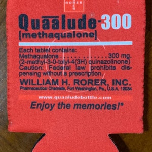 May include: A red fabric beverage insulator with the text "Quaalude-300" and "Enjoy the memories!" printed in white and black. The insulator also includes pharmaceutical information and the website www.quaaludebottle.com.
