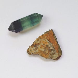 May include: A polished, double-terminated fluorite crystal with green and purple hues, and a piece of rough, orange-brown rock. The crystal is approximately 3 inches long. The objects are set against a white background.