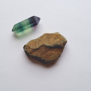 Puede incluir: Una punta de cristal de fluorita verde, morada y transparente junto a una piedra áspera y marrón. El cristal es una punta de doble terminación y la piedra tiene una superficie texturizada. Los objetos están sobre un fondo blanco.