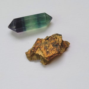 Puede incluir: Un cristal de fluorita pulido de doble terminación, con bandas verdes, moradas y claras. Junto a él, un trozo de roca rugosa, texturizada, marrón y beige. Ambos objetos sobre un fondo blanco.