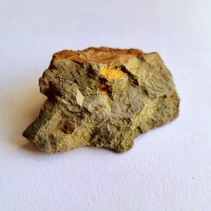 Puede incluir: Una muestra de roca en bruto con una mezcla de colores, incluyendo verde oliva, marrón y amarillo. La roca tiene una superficie texturizada con capas visibles y pequeñas inclusiones de color marrón rojizo. La forma general es irregular y natural.