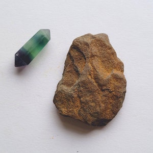 May include: A polished, double-terminated fluorite crystal in shades of green, purple, and clear, next to a rough, brown rock. The crystal is approximately 2 inches long. The rock has a textured surface and irregular shape, with a rusty brown color.