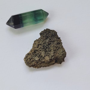 May include: A double-terminated fluorite crystal with green and purple hues, and a piece of rough, dark brown rock. The crystal is a polished gemstone, while the rock has a textured, uneven surface. Both objects are set against a white background.