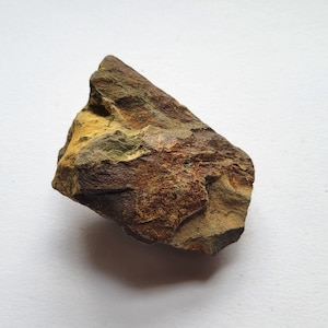 May include: A rough, irregularly shaped rock with a mix of earthy colors. The stone features shades of brown, tan, and hints of green and yellow. The surface has a textured appearance, with visible layers and variations in color.