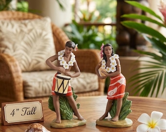Pode incluir: Duas estatuetas de dançarinos com colares de flores brancas e saias laranja. Um dançarino toca um tambor, enquanto o outro faz um gesto de hula. Uma placa indica 4" Tall, com uma concha e uma flor de plumeria branca sobre a superfície.