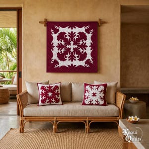 May include: A square wall quilt featuring a deep burgundy background with a white botanical applique pattern, displayed above a light brown rattan sofa with two matching square throw pillows, each showcasing a white floral design on burgundy.
