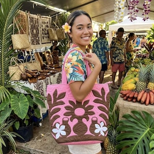 May include: A woman holding a pink tote bag with a brown floral design. The bag is filled with items and the woman is smiling. The background includes tropical plants, fruit, and other market items.