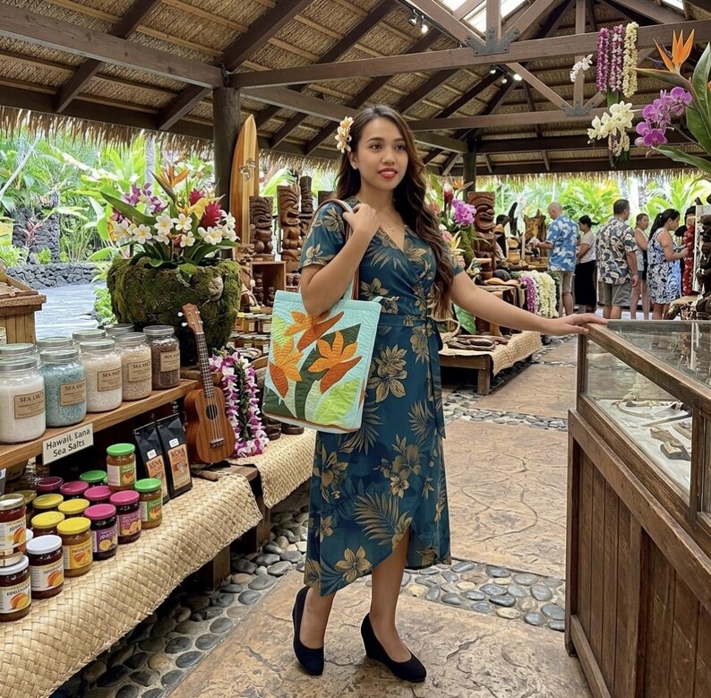 May include: A teal wrap dress with a floral pattern and a matching tote bag featuring orange flowers. The scene is set in a shop with wooden carvings, jars of sea salt, and a ukulele.