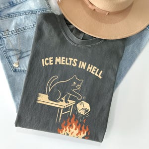 May include: Dark gray t-shirt with the text "ICE MELTS IN HELL" and a graphic of a black cat pushing an ice cube into flames. The shirt is laid on top of blue jeans and a tan hat.