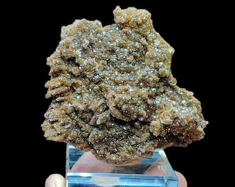 May include: A brown mineral specimen with a crystalline structure, displayed on a clear acrylic stand. The mineral has a rough, textured surface with sparkling, reflective areas. The background is black, highlighting the mineral's details.