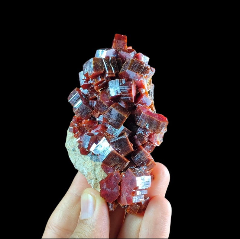May include: A cluster of deep red, translucent vanadinite crystals with a hexagonal, prismatic structure. The mineral specimen features numerous small, sharp-edged crystals densely packed together on a light-colored, textured rocky matrix base.