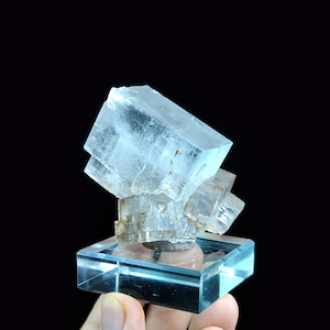 Op de afbeelding: Een helder, kristallijn mineraalexemplaar met kubusvormige structuren en zichtbare interne breuken, gemonteerd op een vierkante, transparante acrylbasis. Het cluster heeft scherpe, geometrische randen en een doorschijnend uiterlijk tegen een donkere achtergrond.