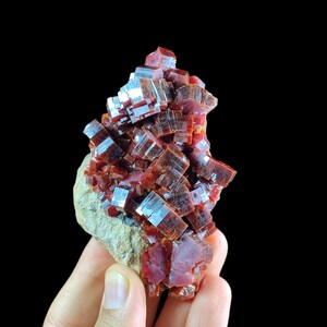 May include: A cluster of deep red, translucent vanadinite crystals with a hexagonal, prismatic structure. The mineral formation features sharp, blocky edges and a glossy, reflective surface, attached to a light tan, textured rocky matrix base.