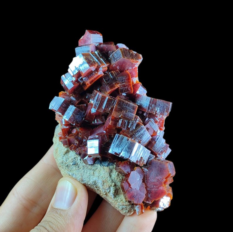 May include: A cluster of deep red, translucent vanadinite crystals with a hexagonal, prismatic structure. The mineral formation is attached to a rough, light brown, earthy matrix base, showcasing a high-gloss, reflective surface texture.