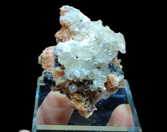 May include: A mineral specimen featuring a cluster of translucent, crystalline formations atop a base of orange and black minerals. The specimen is displayed on a clear, rectangular stand against a black background.