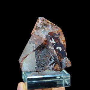 May include: A clear quartz crystal with a reddish-brown inclusion. The crystal is on a clear glass display stand.