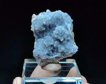 May include: A cluster of light blue crystals with a sparkling texture. The crystal formation is mounted on a clear, rectangular display stand. The background is black.