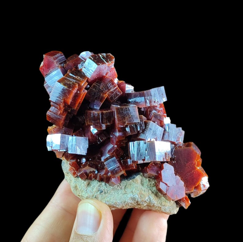 May include: A cluster of deep red, translucent vanadinite crystals with a hexagonal, tabular structure. The mineral specimen features a glossy, reflective surface and is attached to a light gray, textured, rocky matrix base.