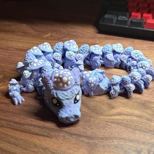 May include: A light blue articulated dragon figurine with mushroom-shaped details and white polka dots. The dragon is displayed on a wooden surface, with a keyboard in the background. The figurine is a unique decorative item.