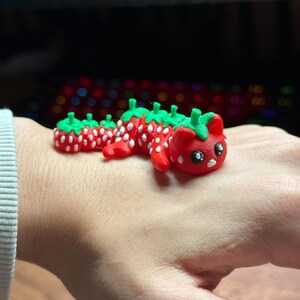 May include: A red and green clay figurine of a cat with a strawberry design. The cat is laying on its belly with its head turned to the side.