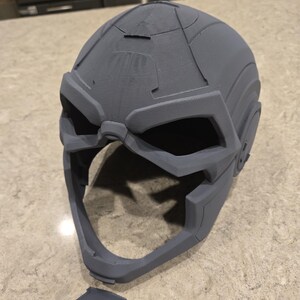 May include: A gray, 3D-printed helmet with a futuristic design. The helmet features angular shapes, large eye openings, and a prominent chin area. A detached piece lies below the helmet.