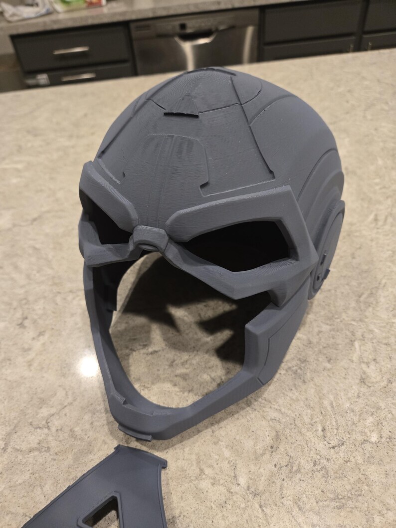 May include: A gray, 3D-printed helmet with a futuristic design. The helmet features angular shapes, large eye openings, and a prominent chin area. A detached piece lies below the helmet.