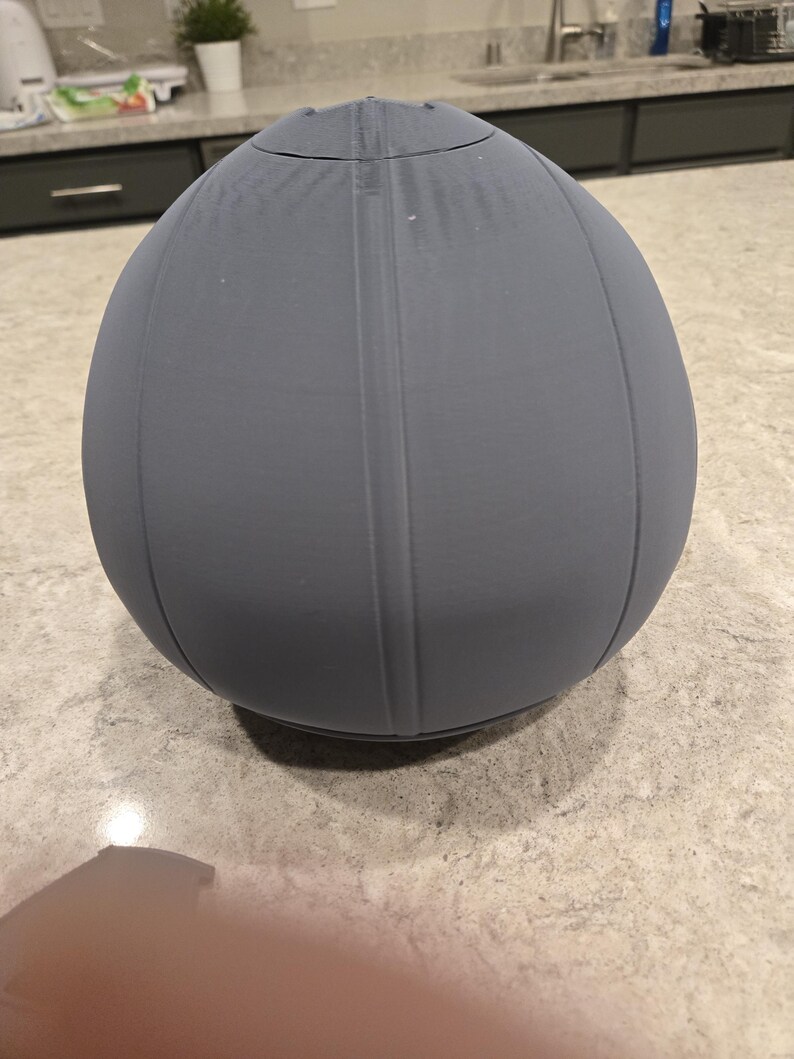 May include: A gray, egg-shaped object with vertical lines and a rounded top. The object appears to be made of a solid material and has a smooth, matte finish. The object is sitting on a speckled surface.
