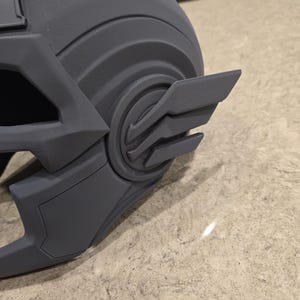 May include: A gray, 3D-printed helmet with a detailed design. The helmet features a prominent wing-like emblem on the side and a curved, ridged structure. The overall aesthetic suggests a futuristic or fantasy theme.