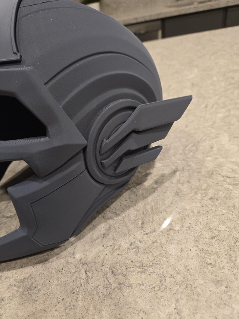 May include: A gray, 3D-printed helmet with a detailed design. The helmet features a prominent wing-like emblem on the side and a curved, ridged structure. The overall aesthetic suggests a futuristic or fantasy theme.