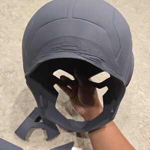 May include: Gray helmet with a face guard and separate wing and letter "A" components. The helmet has a textured surface and appears to be 3D-printed. The design suggests a superhero or cosplay theme.