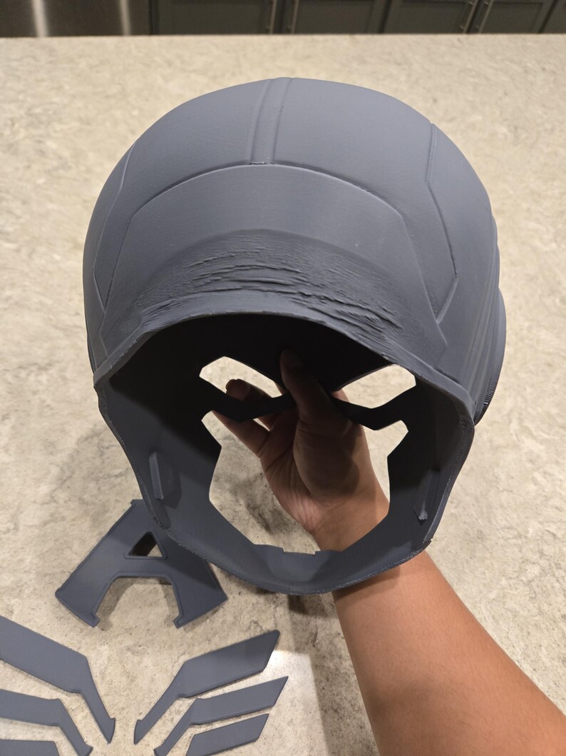 May include: Gray helmet with a face guard and separate wing and letter "A" components. The helmet has a textured surface and appears to be 3D-printed. The design suggests a superhero or cosplay theme.