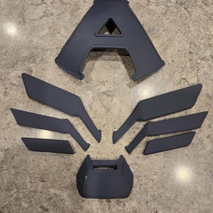 May include: A collection of dark gray 3D-printed components, including a large letter "A", wing-like shapes, and other geometric pieces. The parts are arranged on a speckled gray surface, suggesting a construction or assembly project.