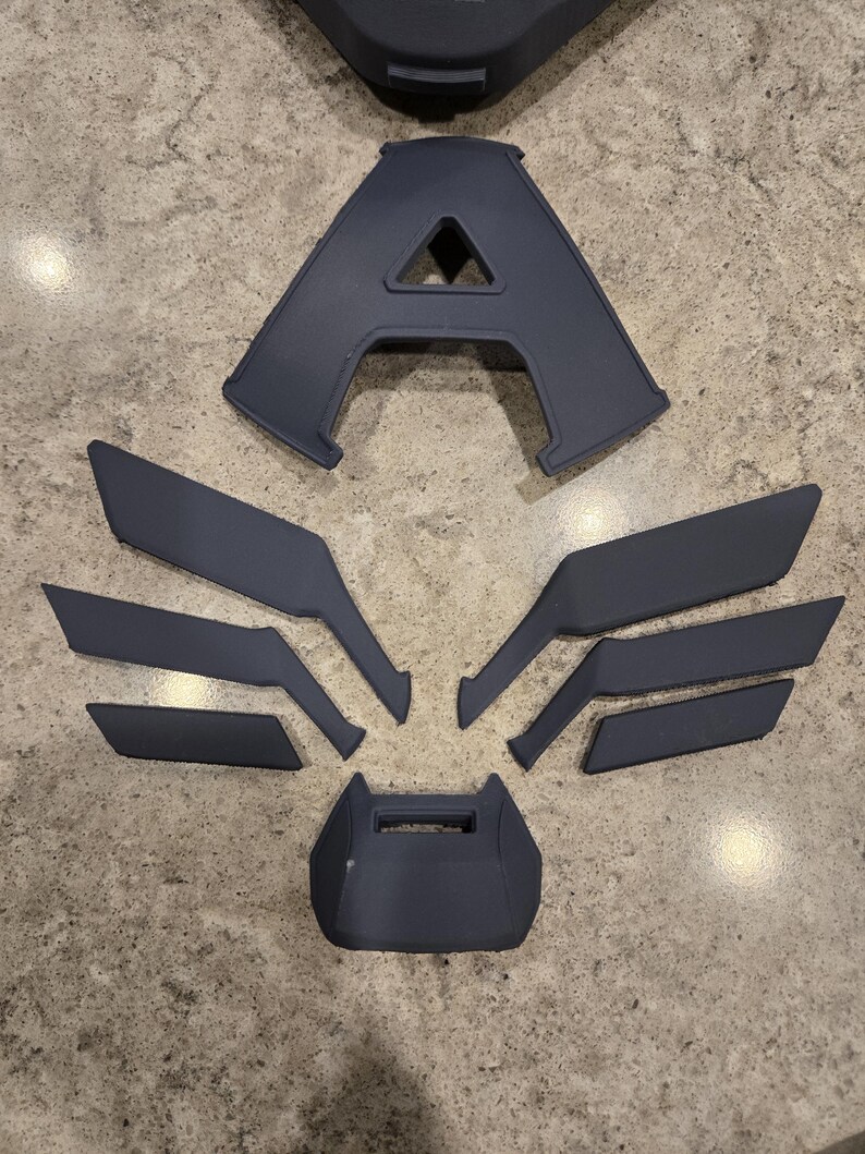May include: A collection of dark gray 3D-printed components, including a large letter "A", wing-like shapes, and other geometric pieces. The parts are arranged on a speckled gray surface, suggesting a construction or assembly project.