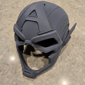 May include: A gray, 3D-printed helmet with an "A" emblem on the forehead. The helmet features a visor and ear coverings, and is likely a cosplay or costume piece. The design is detailed and appears to be a replica.