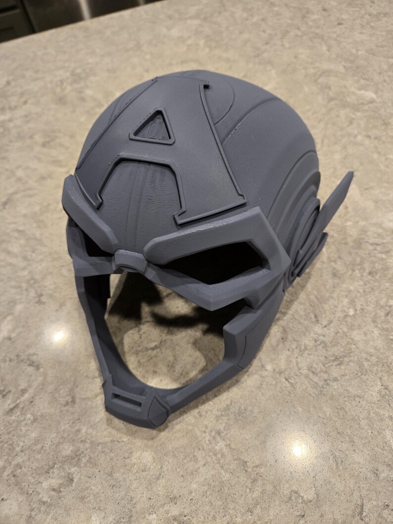 May include: A gray, 3D-printed helmet with an "A" emblem on the forehead. The helmet features a visor and ear coverings, and is likely a cosplay or costume piece. The design is detailed and appears to be a replica.