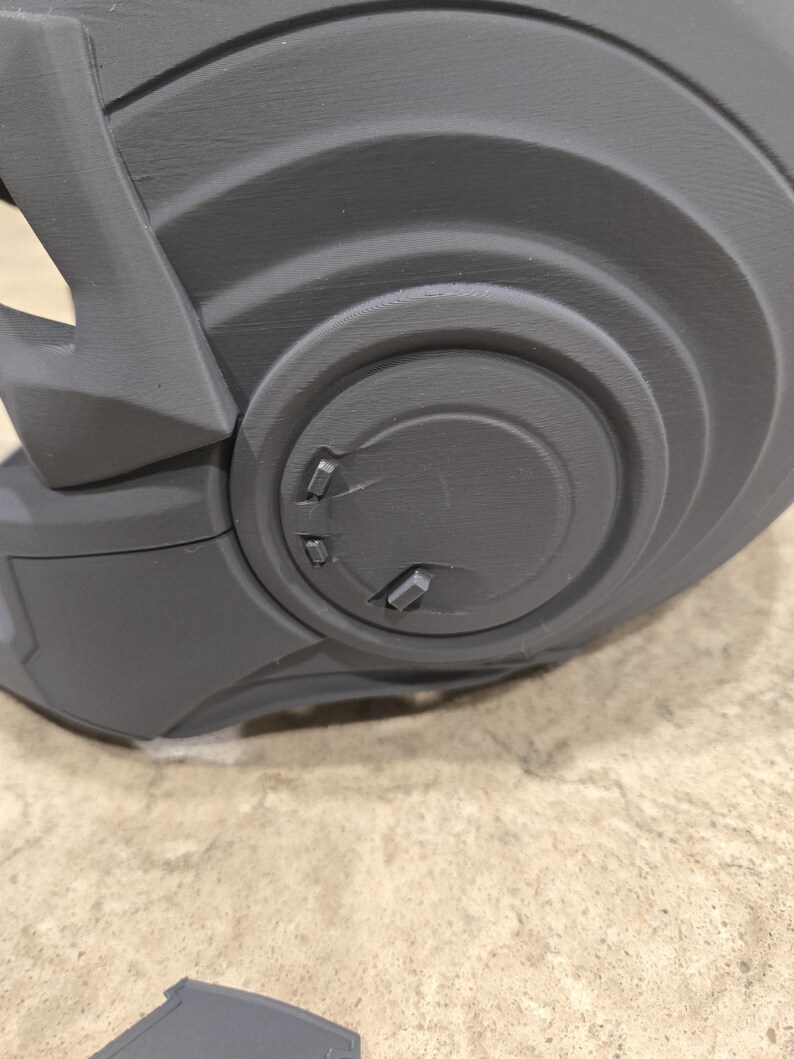 May include: Close-up of a gray, 3D-printed helmet component. The component features a series of concentric circular designs with raised details and small, rectangular protrusions. The surface has a textured appearance.