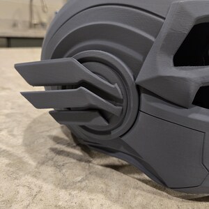 May include: A close-up of a gray, angular helmet with intricate details. The helmet features a series of layered, wing-like structures on the side and a textured surface. The design suggests a futuristic or sci-fi theme.