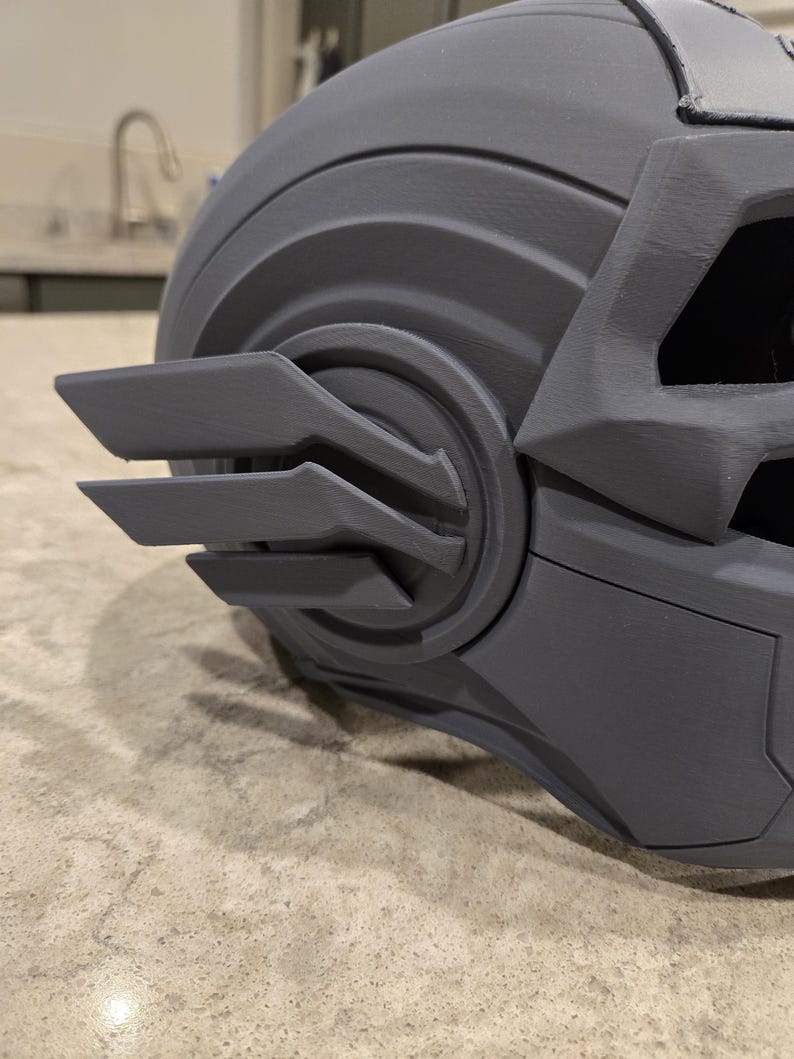 May include: A close-up of a gray, angular helmet with intricate details. The helmet features a series of layered, wing-like structures on the side and a textured surface. The design suggests a futuristic or sci-fi theme.